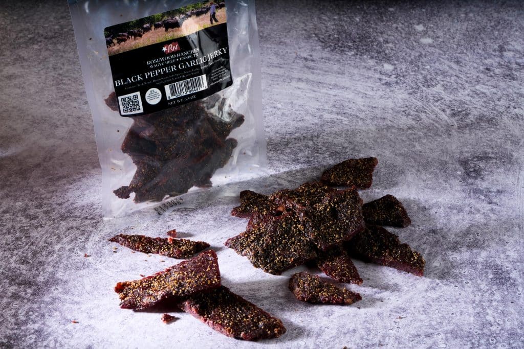 Black Pepper Garlic Jerky from Rosewood Beef, showcasing pieces of jerky scattered around a resealable pouch, emphasizing premium beef quality.