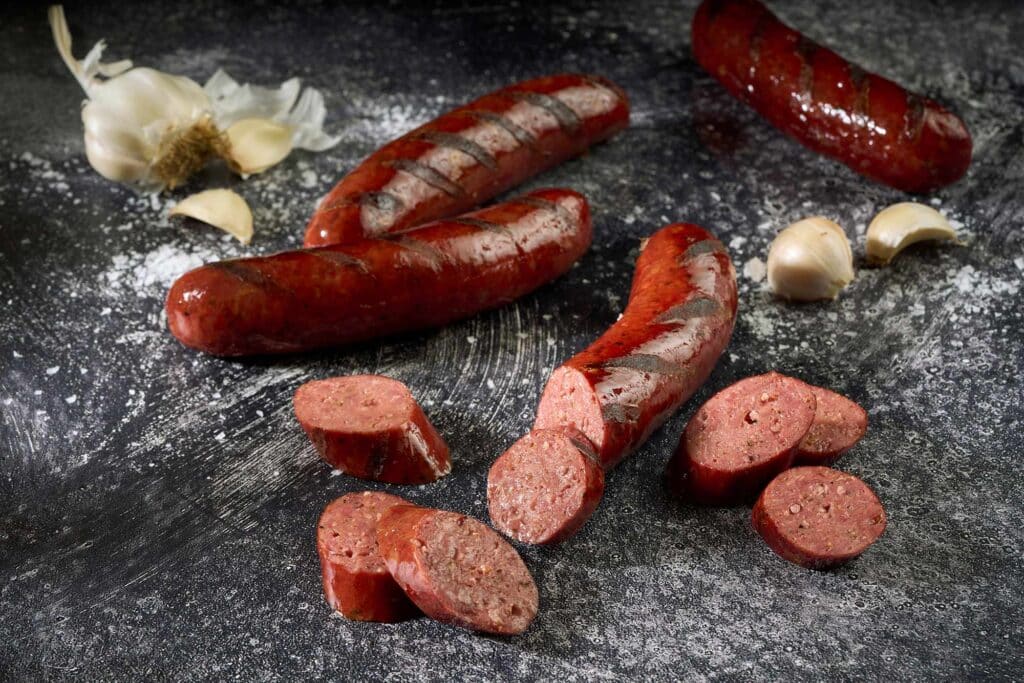Hickory-smoked Wagyu sausage links and sliced pieces on a dark surface, accompanied by garlic cloves, showcasing a bold and decadent product from Rosewood Beef.