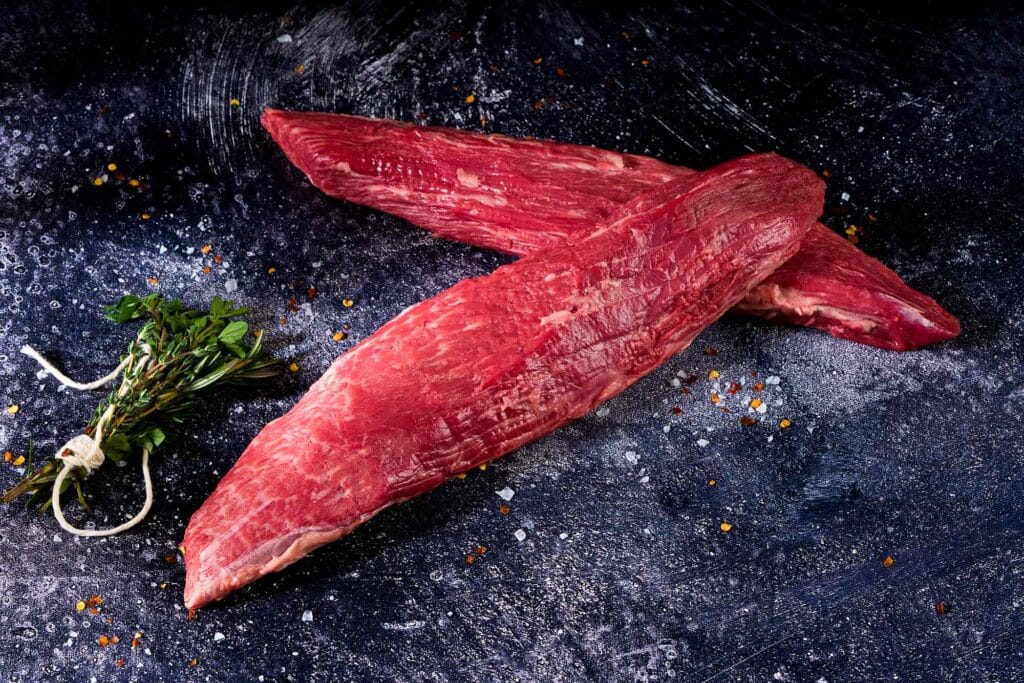 Wagyu Chuck Tender cut showcasing dense structure and light marbling, accompanied by fresh herbs on a textured dark surface, emphasizing culinary elegance and versatility.