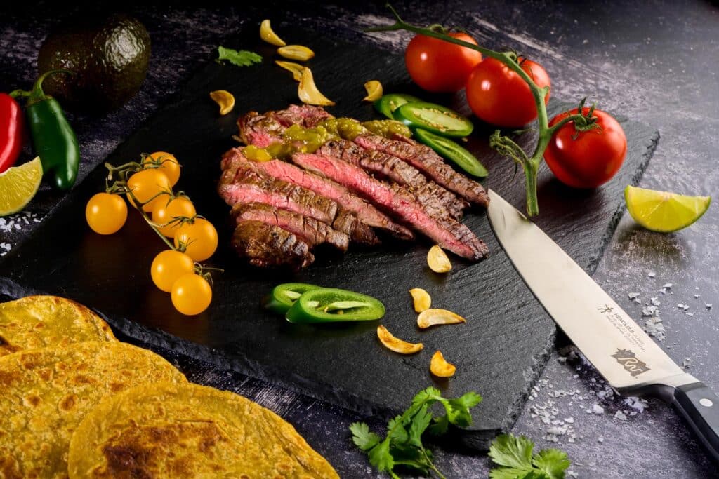 Wagyu outside skirt steak sliced and garnished with fresh vegetables, including tomatoes, jalape&ntilde;os, and yellow cherry tomatoes, on a slate board, accompanied by tortillas and a knife, showcasing premium beef quality.