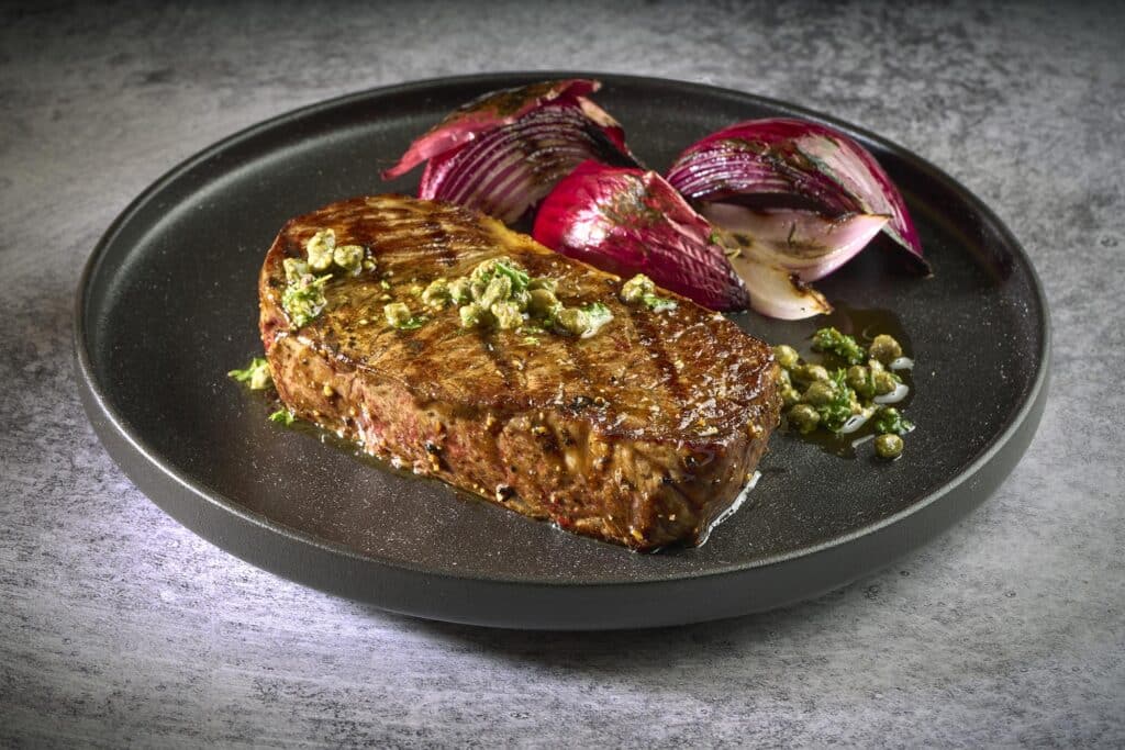 Wagyu strip loin steak garnished with herbs, accompanied by grilled red onions on a black plate, showcasing premium beef quality from Rosewood Beef.