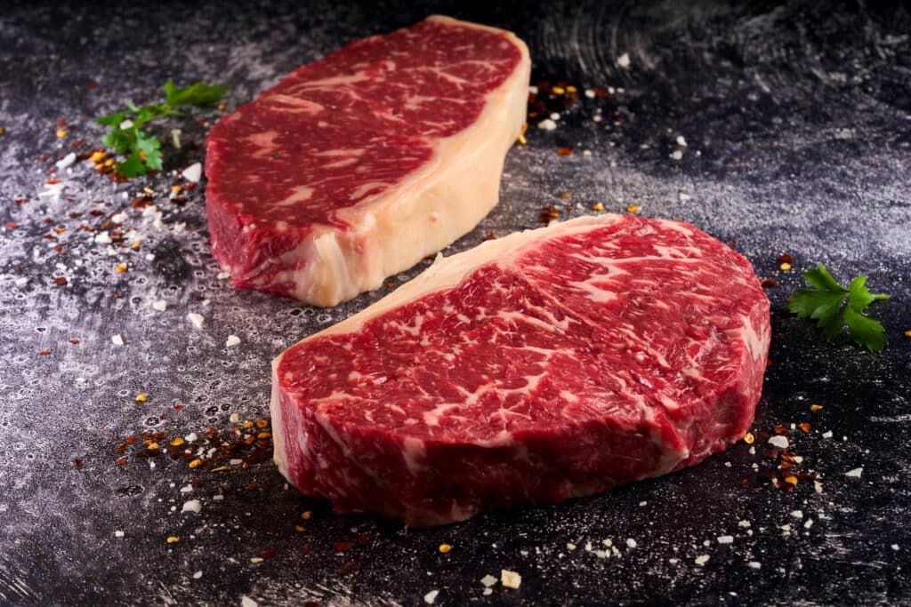 Wagyu Strip Loin steaks showcasing deep marbling and rich fat cap, positioned on a dark surface with seasoning and herbs, emphasizing premium quality and culinary elegance.