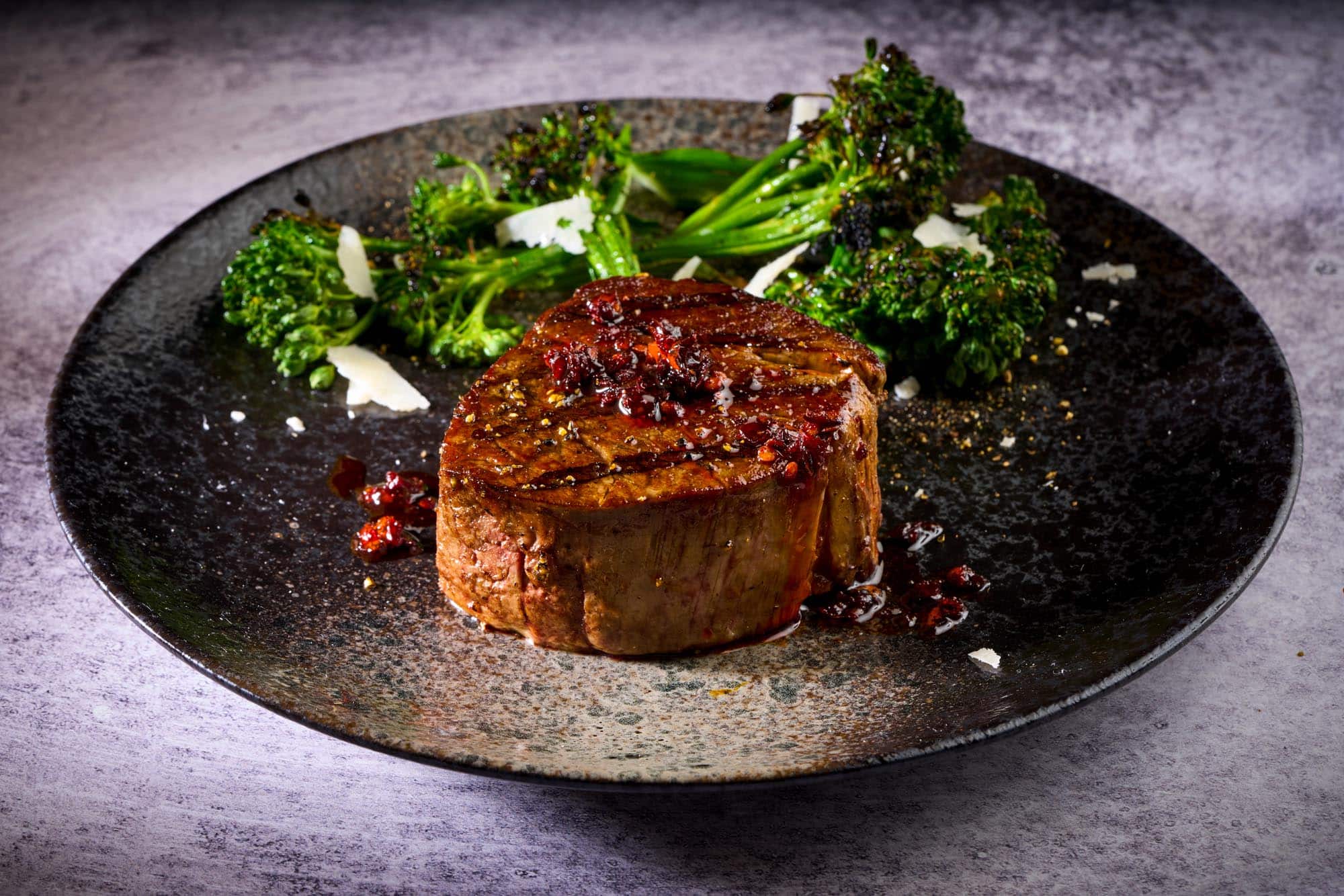 Wagyu tenderloin steak garnished with herbs and seasoning, served with broccolini on a textured black plate, highlighting premium quality for culinary enthusiasts.