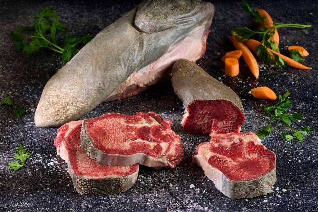 Boneless Wagyu tongue cut into slices showcasing rich marbling and texture, with a whole tongue piece in the background, garnished with fresh parsley and carrots, emphasizing culinary versatility and depth of flavor.