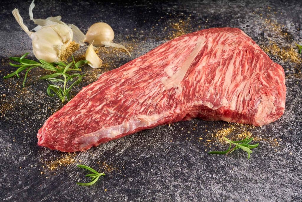 Wagyu Tri Tip cut with elegant marbling and tight grain, garnished with garlic and fresh herbs on a textured surface.
