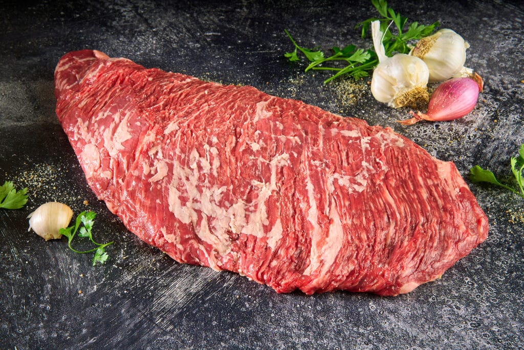 Wagyu Sirloin Flap cut with rich marbling and loose grain, surrounded by garlic, shallots, and parsley on a dark surface, highlighting its premium quality and flavor profile for culinary applications.